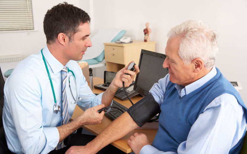 GP doctor taking senior man's blood pressure in surgery room having a check up.