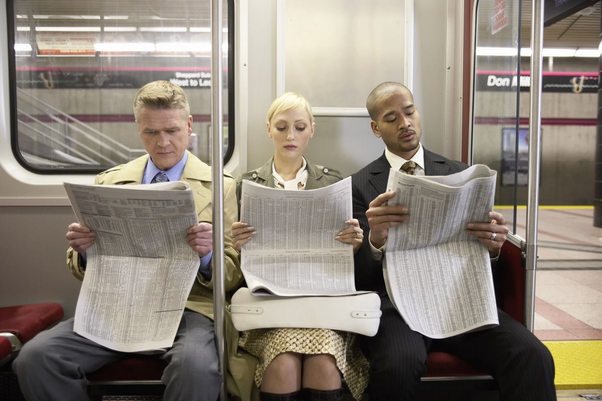 Three people on a subway train read newspapers. ​