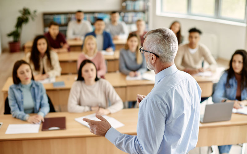 Back view of mature professor giving lecture to large group of college students in the classroom.