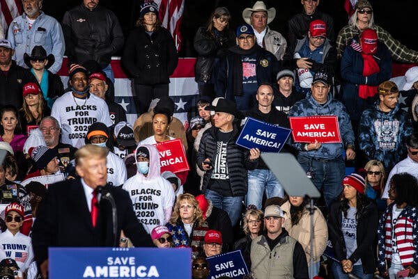 Donald J. Trump stands at a podium at a rally as his supporters stand behind him.