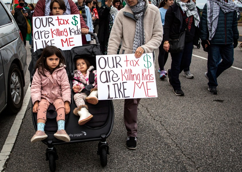 ARLINGTON, VA, UNITED STATES - 2023/12/09: Parents with their kids holding a sign with Our tax dollar are killing kids like me written on it as they join in a demonstration. Pro-Palestinian demonstrators gather at Pentagon City Metro in Arlington and then march to the City with Palestinian flags and banners, calling for a permanent ceasefire in Gaza. (Photo by Probal Rashid/LightRocket via Getty Images)