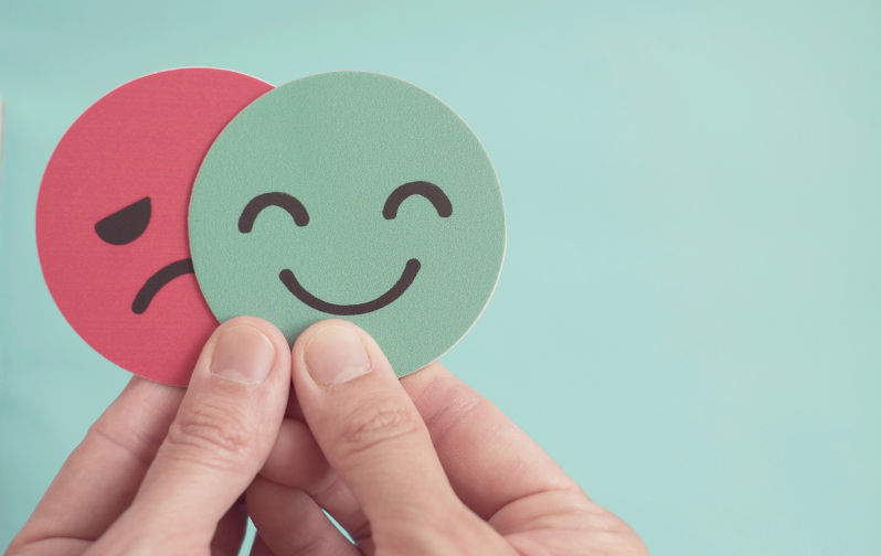 Mood-change. Image: iStock / :ThitareeSarmkasat Hands holding sad face hiding behind happy face, bipolar and depression, mental health, split personality, mood change,