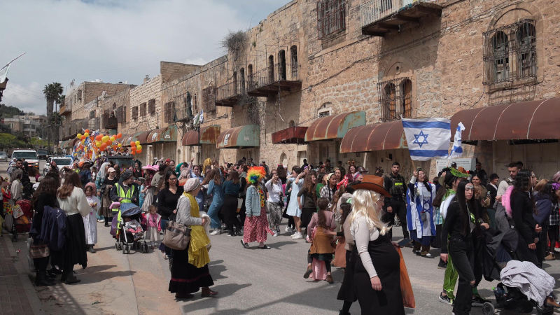 Jewish settlers wearing costumes dancing as they march down Al-Shuhada street, which is largely closed to Palestinians, during the annual Purim parade on March 24, 2024 in Hebron, Israel. Image: Alamy/ Eddie Gerald / Alamy Stock Photo
