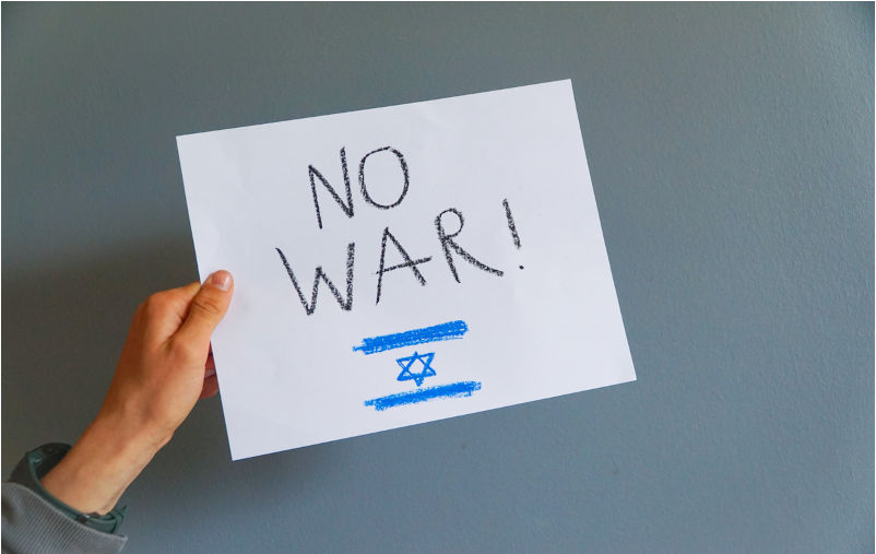 A hand holds up a white sign reading no war, with the Israeli flag as the backdrop, signalling a plea for peace within a national context.